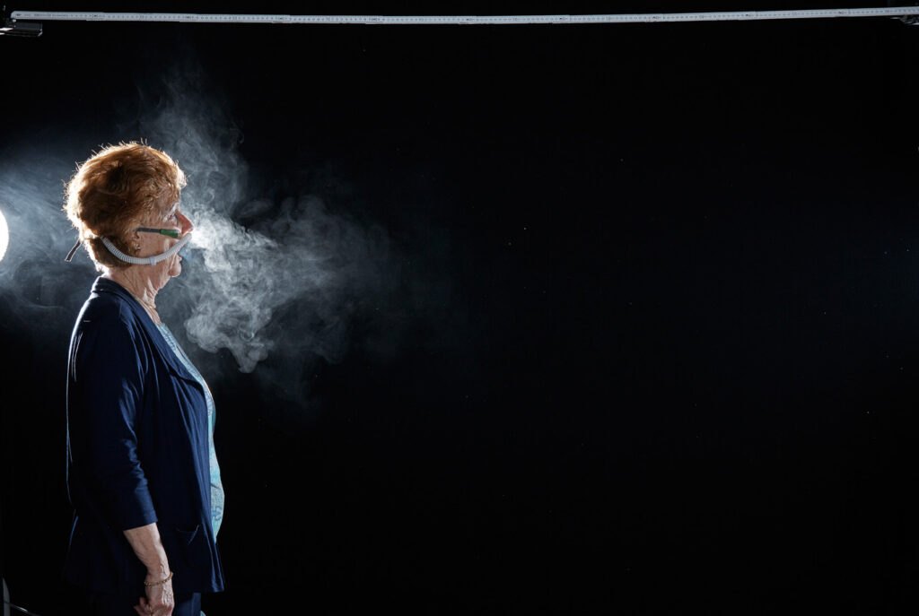 Mischa Nawrata, Fotograf, Aerosolausstoßtest, Chorverband Österreich, Aerosolausstoßtestung, Prof. Dr. Fritz Sterz, Ao.Univ.-Prof. Dr.med.univ. Fritz Sterz, fritz.sterz@meduniwien.ac.at , MedUni Wien, Universitätskliniken der MedUni Wien / AKH Wien, ao.Univ.Prof. Dr. Harald Herkner, MSc, Dr. Heinz Bixa, DDr. Karl-Gerhard Straßl MAS, Präsident, Chorverband Österreich, Mit 4-8 Liter Sauerstoff pro Minute wurde mit einer Düse in einem kleinen Behältnis mit 0,9% Kochsalz Nebel (= Aerosol) erzeugt. Mit kleinen Sonden in den Nasenöffnungen wurde den Sängern dieser Nebel kontinuierlich zusätzlich zum Atmen verabreicht. Die Verstreuung des Aerosols in den oberen Atemwegen war damit gewährleistet. In einem schwarz ausgekleideten Raum befanden sich hinter dem jeweiligen Musiker links und rechts besonders starke Scheinwerfer, um das Ausströmen der Aerosole sichtbar zu machen. Dadurch war es möglich, die Verteilung der Ausatmungsluft im Gegenlicht zu visualisieren und fotographisch zu dokumentieren, ChVOe=Chorverband Österreich, Alt, Tenor, Bass, oM=ohne Maske, mM=mit Maske, mG=mit Gesichtsschutz, mMprivat=private Schutzmaske aus Stoff genäht, FFP2 Schutzmaske, es wurden Vokale und Konsonanten gesungen,