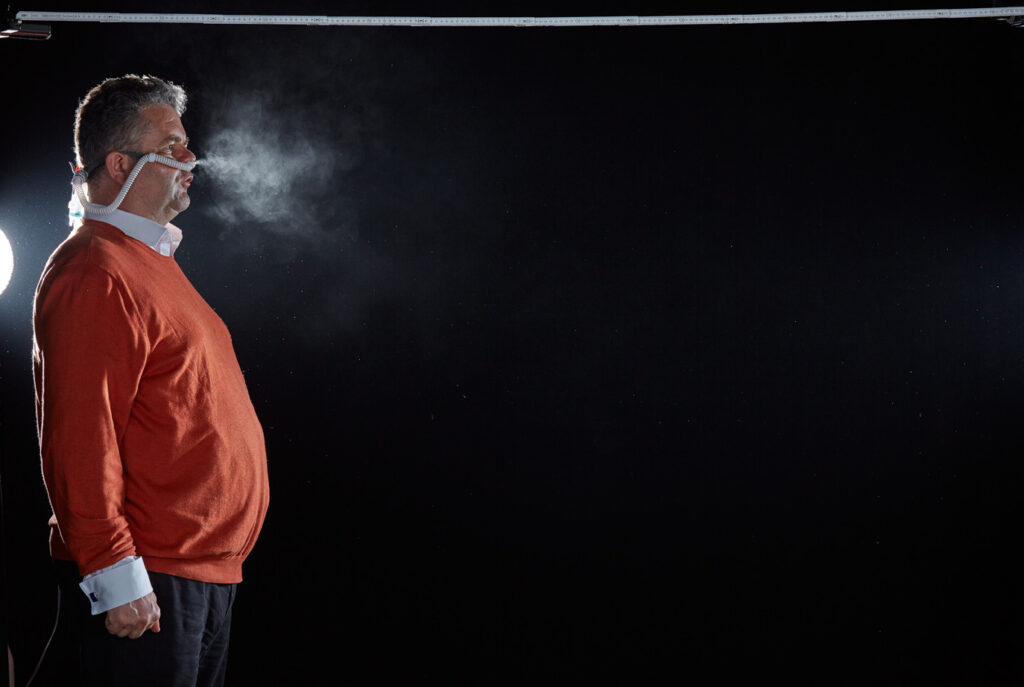 Mischa Nawrata, Fotograf, Aerosolausstoßtest, Chorverband Österreich, Aerosolausstoßtestung, Prof. Dr. Fritz Sterz, Ao.Univ.-Prof. Dr.med.univ. Fritz Sterz, fritz.sterz@meduniwien.ac.at , MedUni Wien, Universitätskliniken der MedUni Wien / AKH Wien, ao.Univ.Prof. Dr. Harald Herkner, MSc, Dr. Heinz Bixa, DDr. Karl-Gerhard Straßl MAS, Präsident, Chorverband Österreich, Mit 4-8 Liter Sauerstoff pro Minute wurde mit einer Düse in einem kleinen Behältnis mit 0,9% Kochsalz Nebel (= Aerosol) erzeugt. Mit kleinen Sonden in den Nasenöffnungen wurde den Sängern dieser Nebel kontinuierlich zusätzlich zum Atmen verabreicht. Die Verstreuung des Aerosols in den oberen Atemwegen war damit gewährleistet. In einem schwarz ausgekleideten Raum befanden sich hinter dem jeweiligen Musiker links und rechts besonders starke Scheinwerfer, um das Ausströmen der Aerosole sichtbar zu machen. Dadurch war es möglich, die Verteilung der Ausatmungsluft im Gegenlicht zu visualisieren und fotographisch zu dokumentieren, ChVOe=Chorverband Österreich, Alt, Tenor, Bass, oM=ohne Maske, mM=mit Maske, mG=mit Gesichtsschutz, mMprivat=private Schutzmaske aus Stoff genäht, FFP2 Schutzmaske, es wurden Vokale und Konsonanten gesungen,