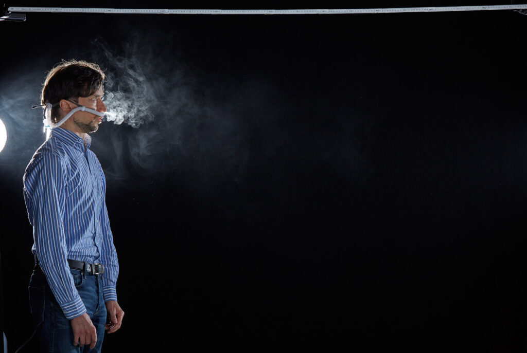 Mischa Nawrata, Fotograf, Aerosolausstoßtest, Chorverband Österreich, Aerosolausstoßtestung, Prof. Dr. Fritz Sterz, Ao.Univ.-Prof. Dr.med.univ. Fritz Sterz, fritz.sterz@meduniwien.ac.at , MedUni Wien, Universitätskliniken der MedUni Wien / AKH Wien, ao.Univ.Prof. Dr. Harald Herkner, MSc, Dr. Heinz Bixa, DDr. Karl-Gerhard Straßl MAS, Präsident, Chorverband Österreich, Mit 4-8 Liter Sauerstoff pro Minute wurde mit einer Düse in einem kleinen Behältnis mit 0,9% Kochsalz Nebel (= Aerosol) erzeugt. Mit kleinen Sonden in den Nasenöffnungen wurde den Sängern dieser Nebel kontinuierlich zusätzlich zum Atmen verabreicht. Die Verstreuung des Aerosols in den oberen Atemwegen war damit gewährleistet. In einem schwarz ausgekleideten Raum befanden sich hinter dem jeweiligen Musiker links und rechts besonders starke Scheinwerfer, um das Ausströmen der Aerosole sichtbar zu machen. Dadurch war es möglich, die Verteilung der Ausatmungsluft im Gegenlicht zu visualisieren und fotographisch zu dokumentieren, ChVOe=Chorverband Österreich, Alt, Tenor, Bass, oM=ohne Maske, mM=mit Maske, mG=mit Gesichtsschutz, mMprivat=private Schutzmaske aus Stoff genäht, FFP2 Schutzmaske, es wurden Vokale und Konsonanten gesungen,